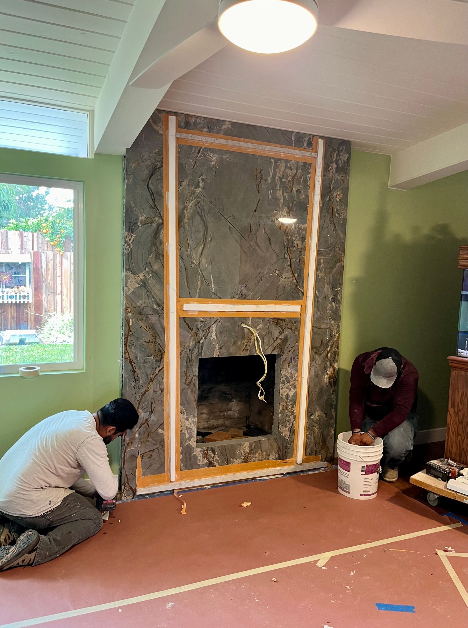 Workers painting around a fireplace wall in a room, with tape, buckets, and a green accent wall.