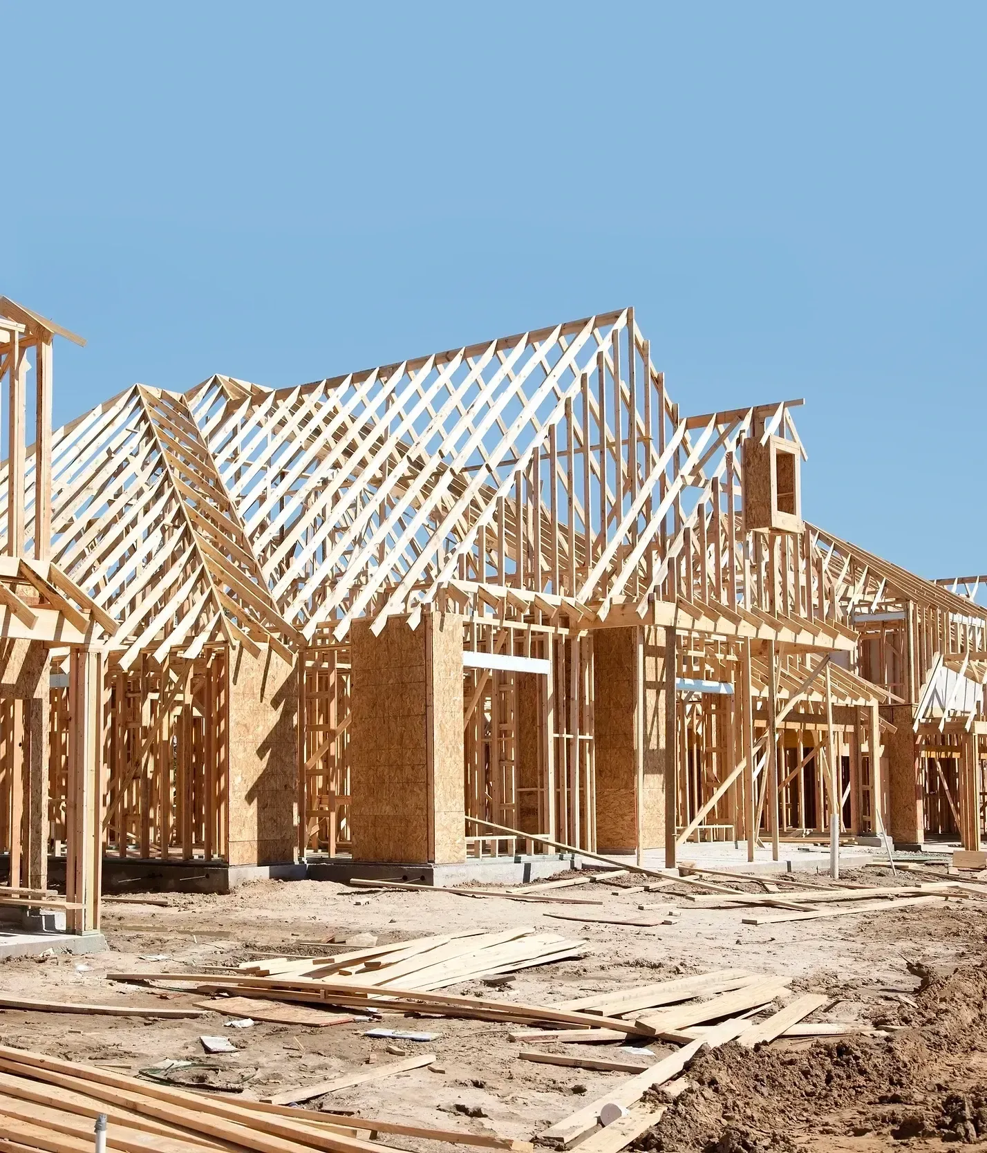 Wood-framed houses under construction on a dusty lot under a clear blue sky