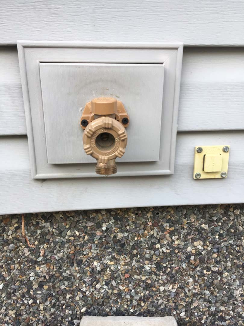 A wall-mounted outdoor water faucet, brass-colored, with a square cover on gray siding.