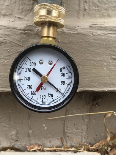 Pressure gauge showing approximately 270 PSI connected to an outdoor spigot on a brick wall.