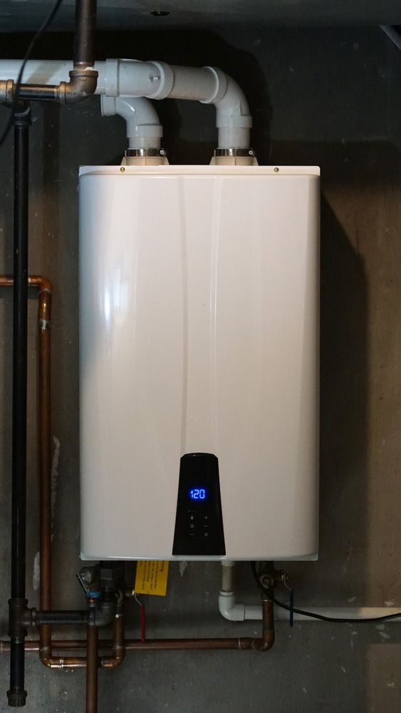 White, wall-mounted tankless water heater with digital display, copper and PVC pipes attached, on a gray wall.