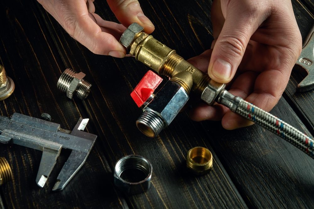 Hands assembling plumbing fittings on a dark wood surface; includes wrench, caliper, and various metal parts.