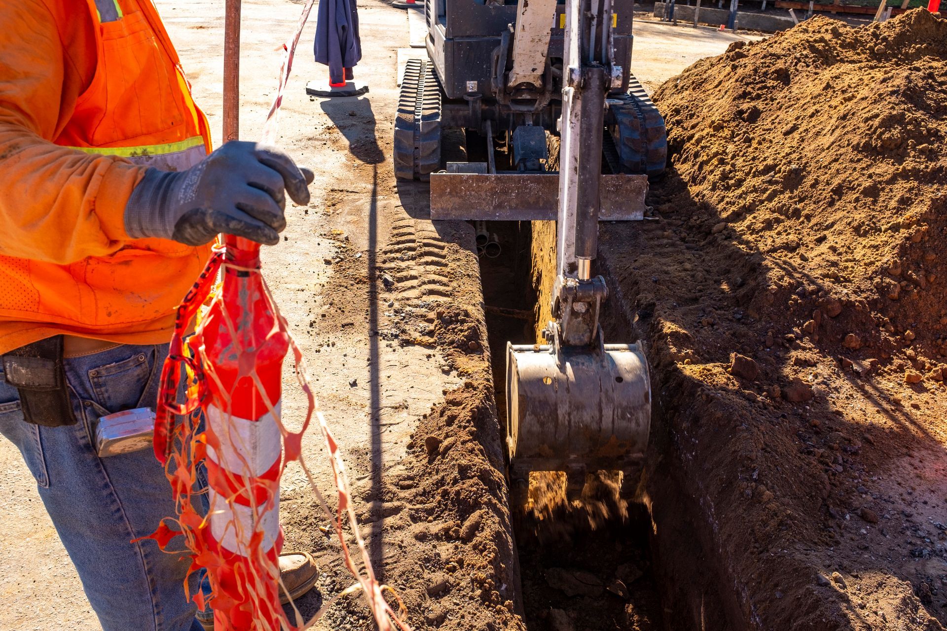 A trenching machine is digging in the street while an unidentified person stands on the left side.