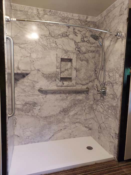 Accessible shower with marble-like walls, grab bars, and built-in shelf. Silver shower fixtures and white base.