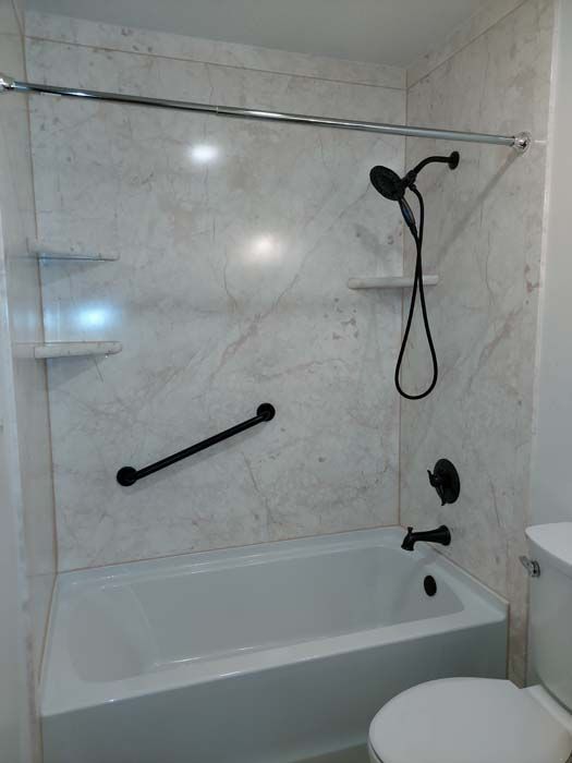 White bathtub and marble-look shower walls with black fixtures and grab bar in a bathroom.
