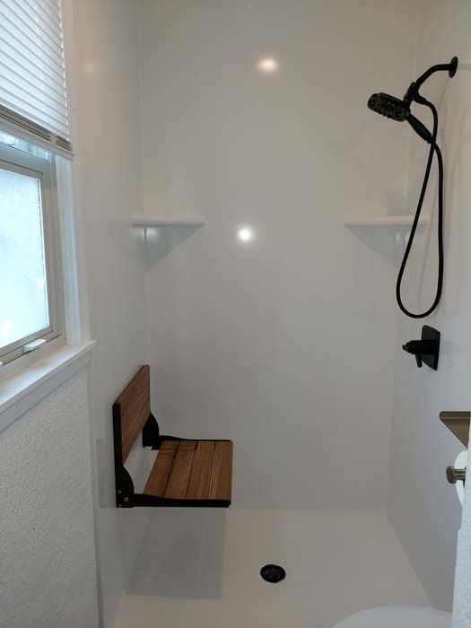 White shower with a foldable wooden seat, black showerhead, and small window.