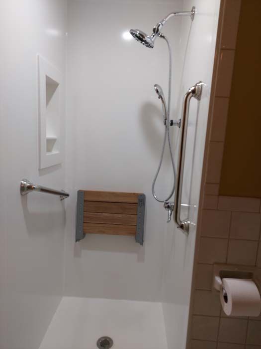 White accessible shower with handrails, a shower chair, and a handheld showerhead.