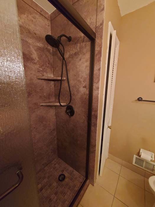 Shower with brown tile walls, black showerhead, and shelves; located next to a door and beige walls.