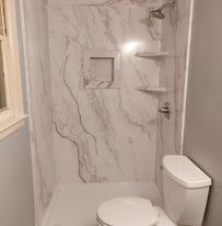 A small bathroom with a white marble-like shower stall, toilet, and shelf.