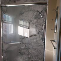 Shower with a marble-patterned wall, glass door, and silver hardware; a window is visible in the reflection.