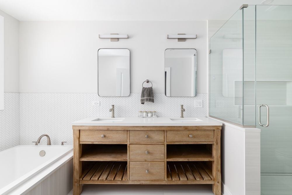 Bathroom with double vanity, mirrors, and shower. Neutral tones, light wood cabinet, white walls, and a bathtub.
