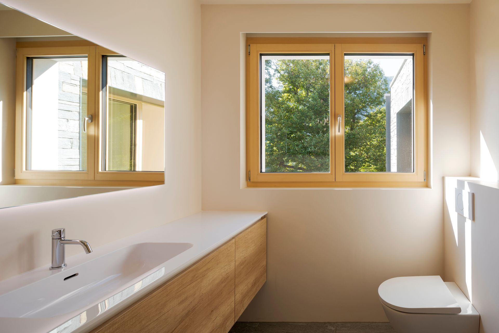Bathroom with wooden-framed window and cabinetry, white sink and toilet; cream walls, sunlight.