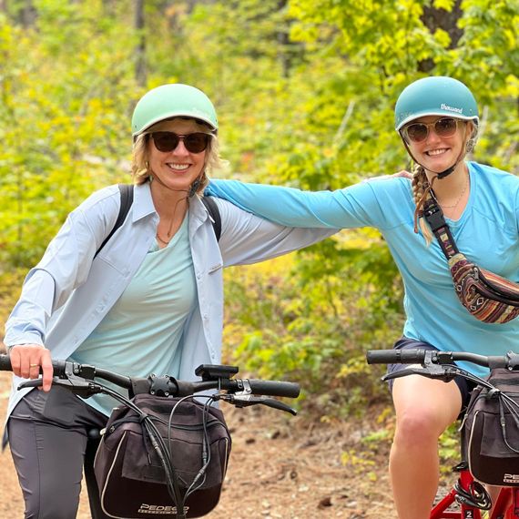 Two women are riding bicycles down a path in the woods.