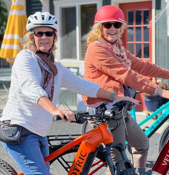 Two women are riding bicycles down a path in the woods.