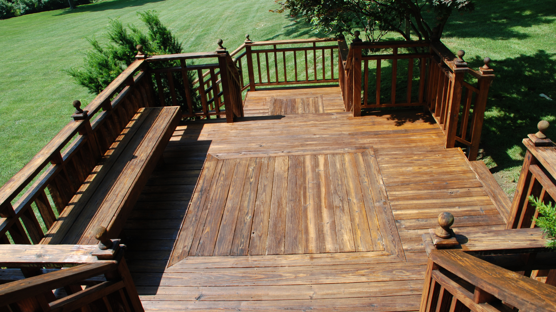 Wooden deck with stairs, built-in bench, and railing; stained brown. Green grass and trees in background.