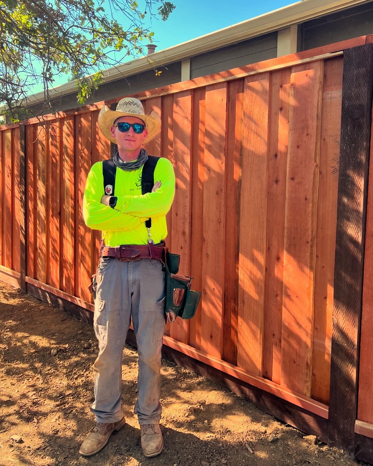 Man in cowboy hat and sunglasses stands in front of a new wooden fence in bright sunlight.