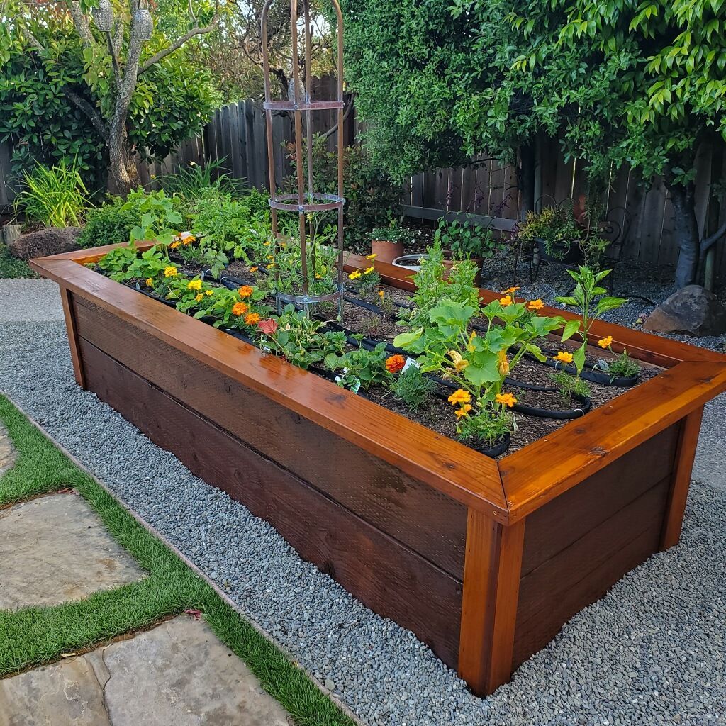 Raised wooden garden bed with flowers and vegetables, set on gravel and next to a patio.