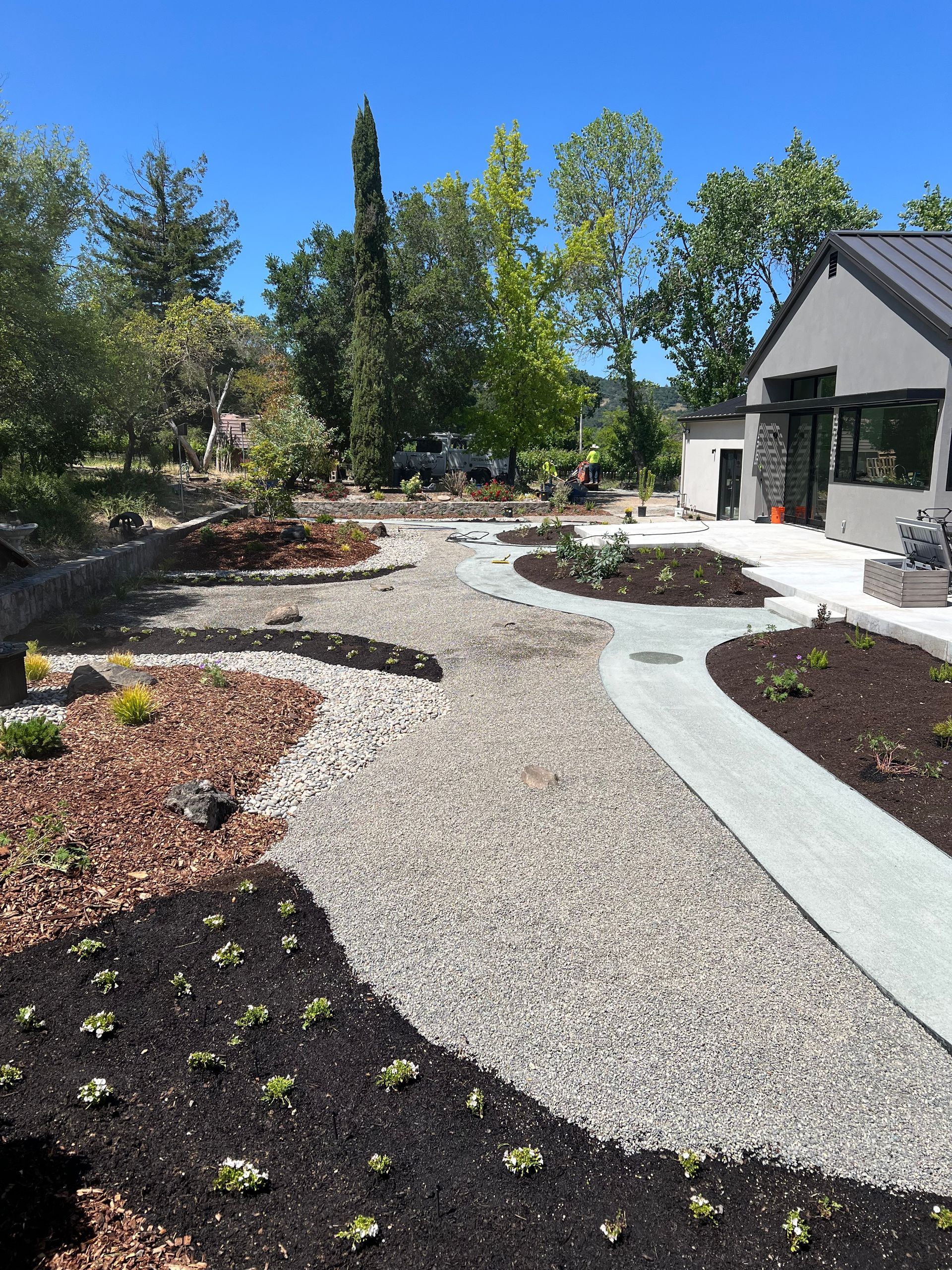 A sunny landscape with gravel paths, flower beds, and a modern house.