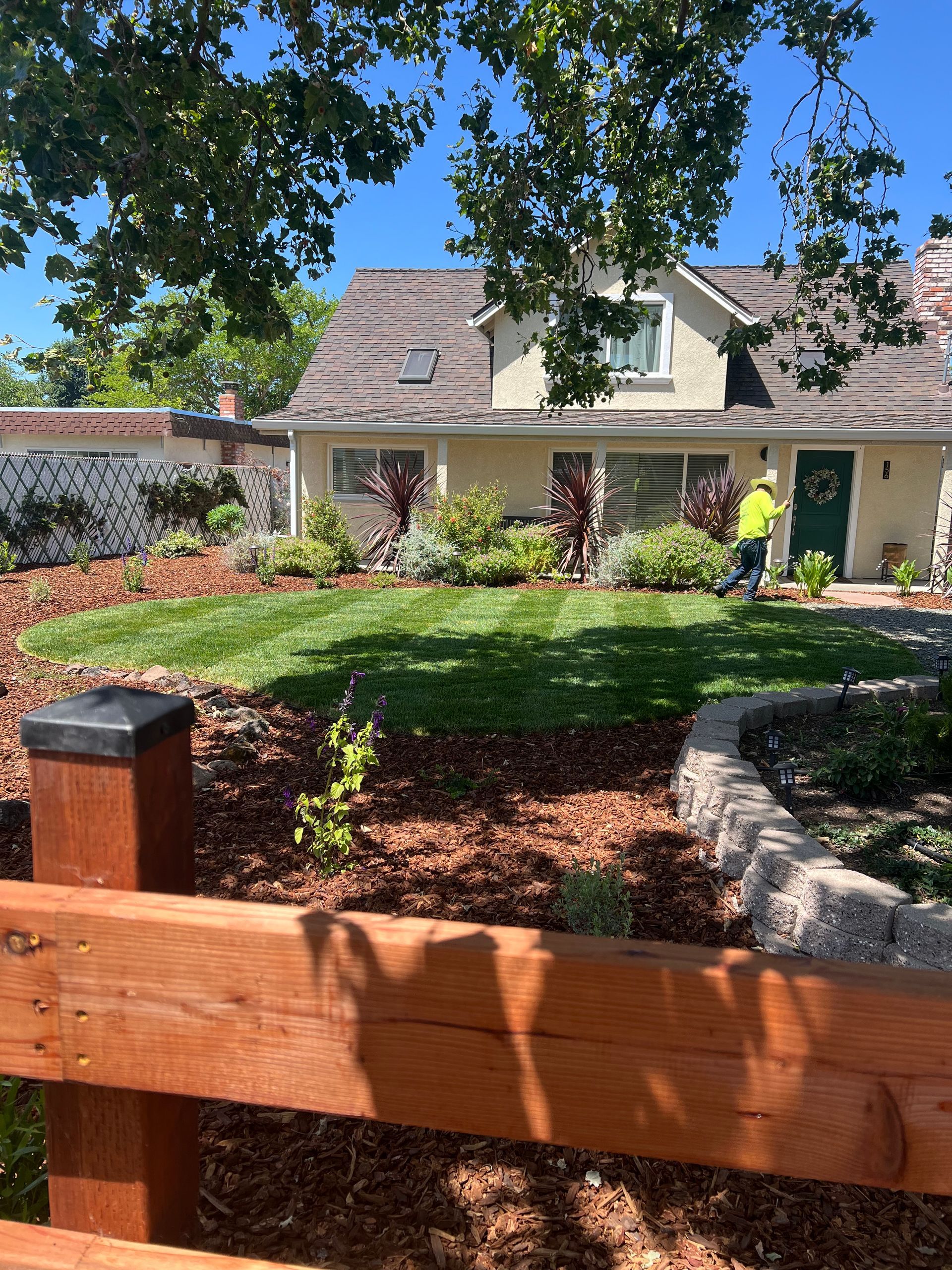 House with fresh lawn, flower beds, and a person mowing; sunny day.