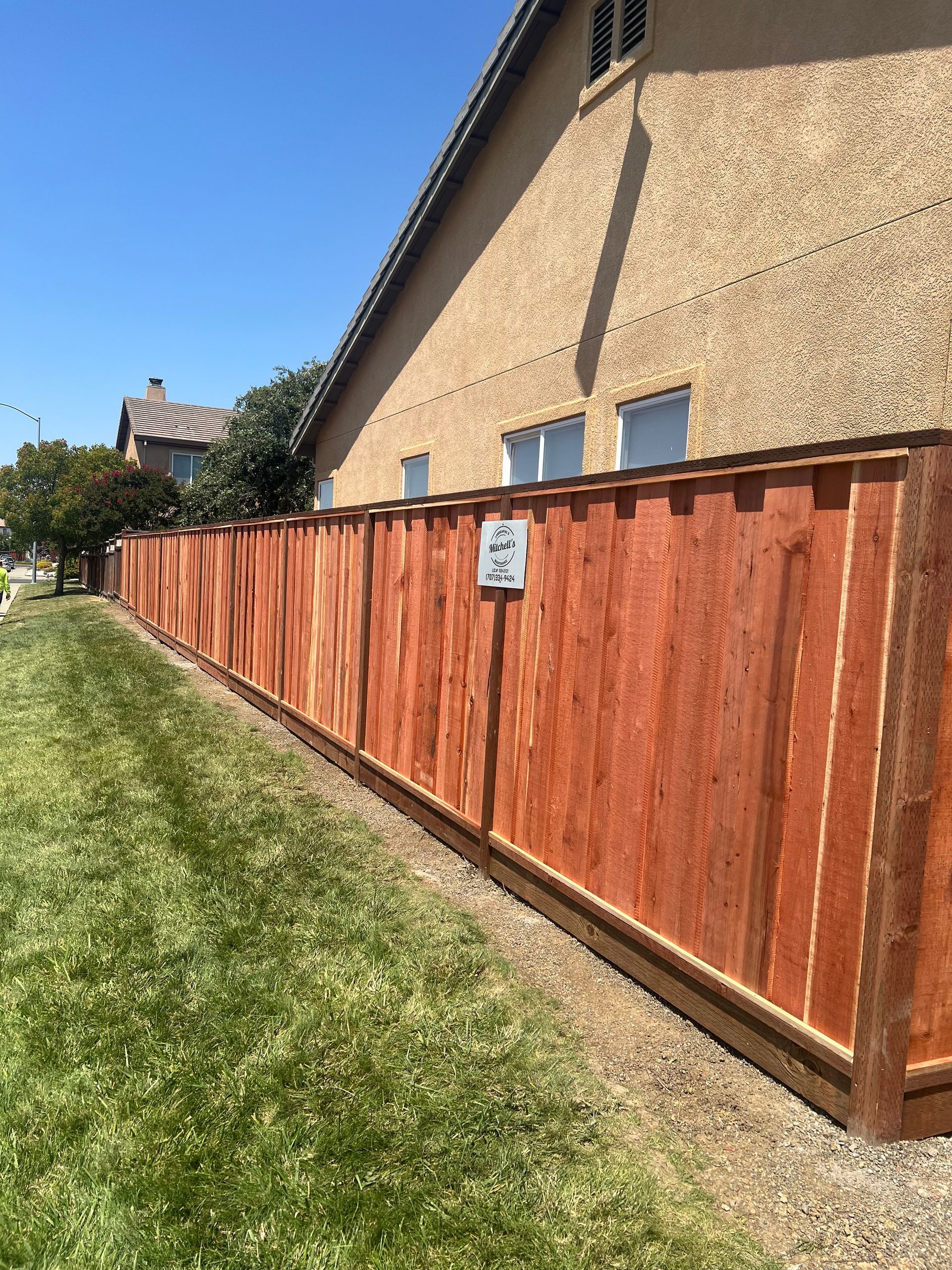 Wooden fence bordering green grass along a tan stucco building under a clear blue sky.