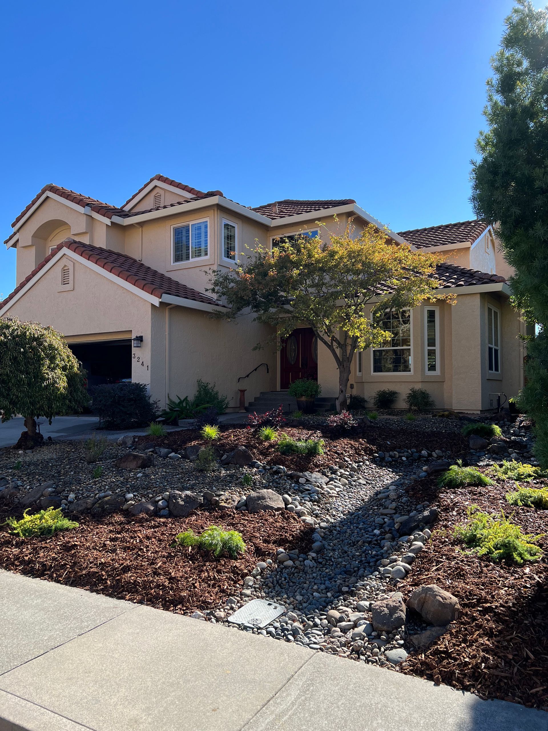 Two-story stucco house with red-tiled roof, beige walls, and dry creek bed landscaping in front.