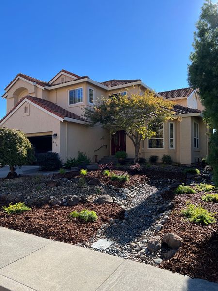 Two-story stucco house with red-tiled roof, beige walls, and dry creek bed landscaping in front.