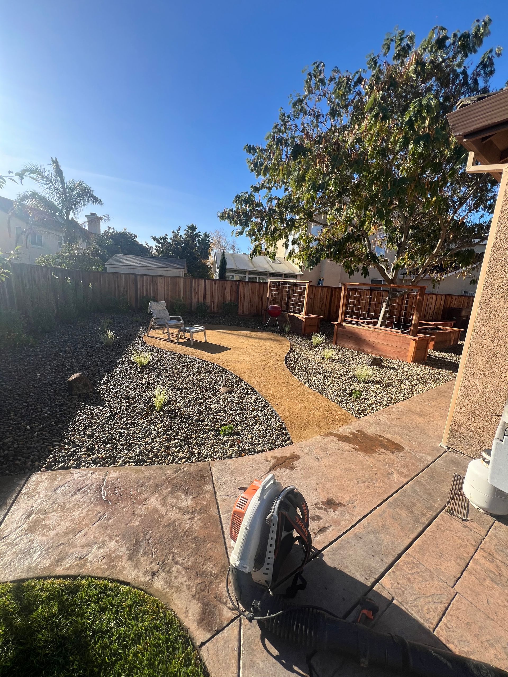 Backyard landscaping with gravel, brick, and a pathway. A leaf blower is in the foreground.