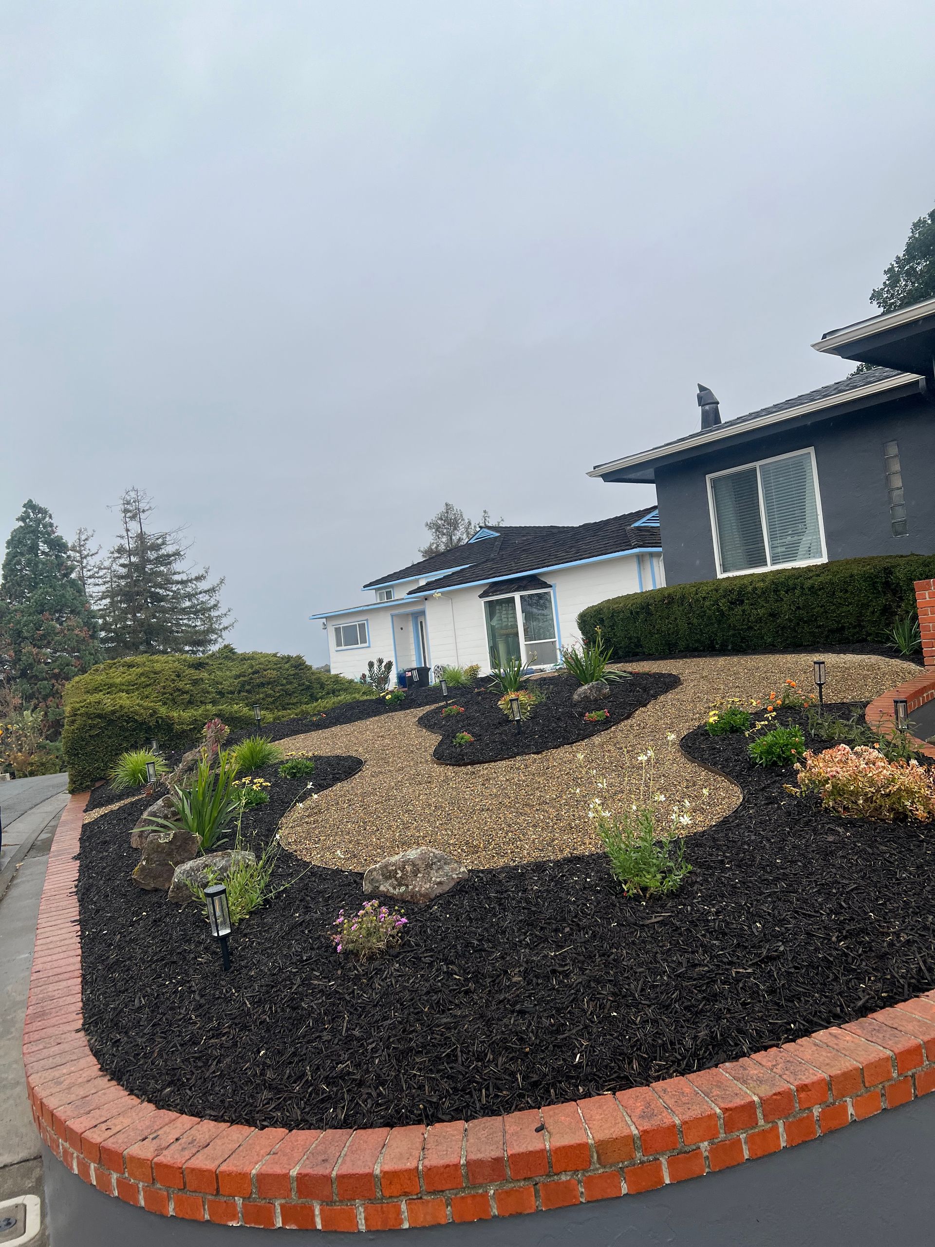 Landscaped yard with dark mulch, tan gravel, and plants in front of a gray house. Cloudy sky.