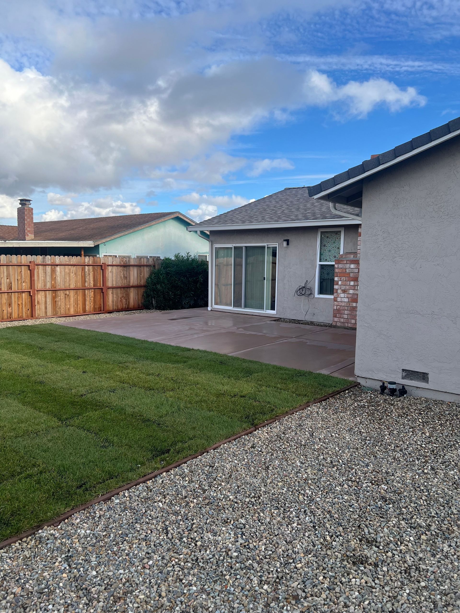 Backyard with grass, patio, gravel, and wooden fence under a cloudy sky.