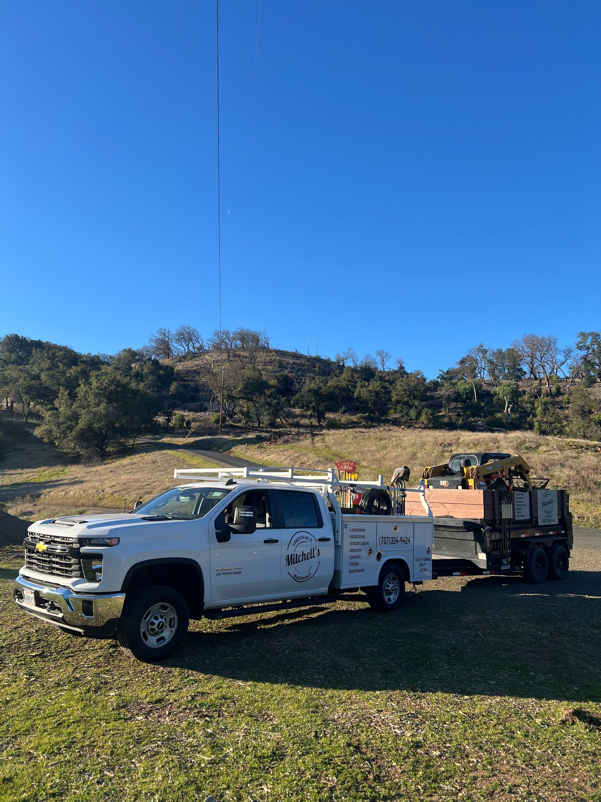 White utility truck towing a trailer on a grassy hill with a clear blue sky.