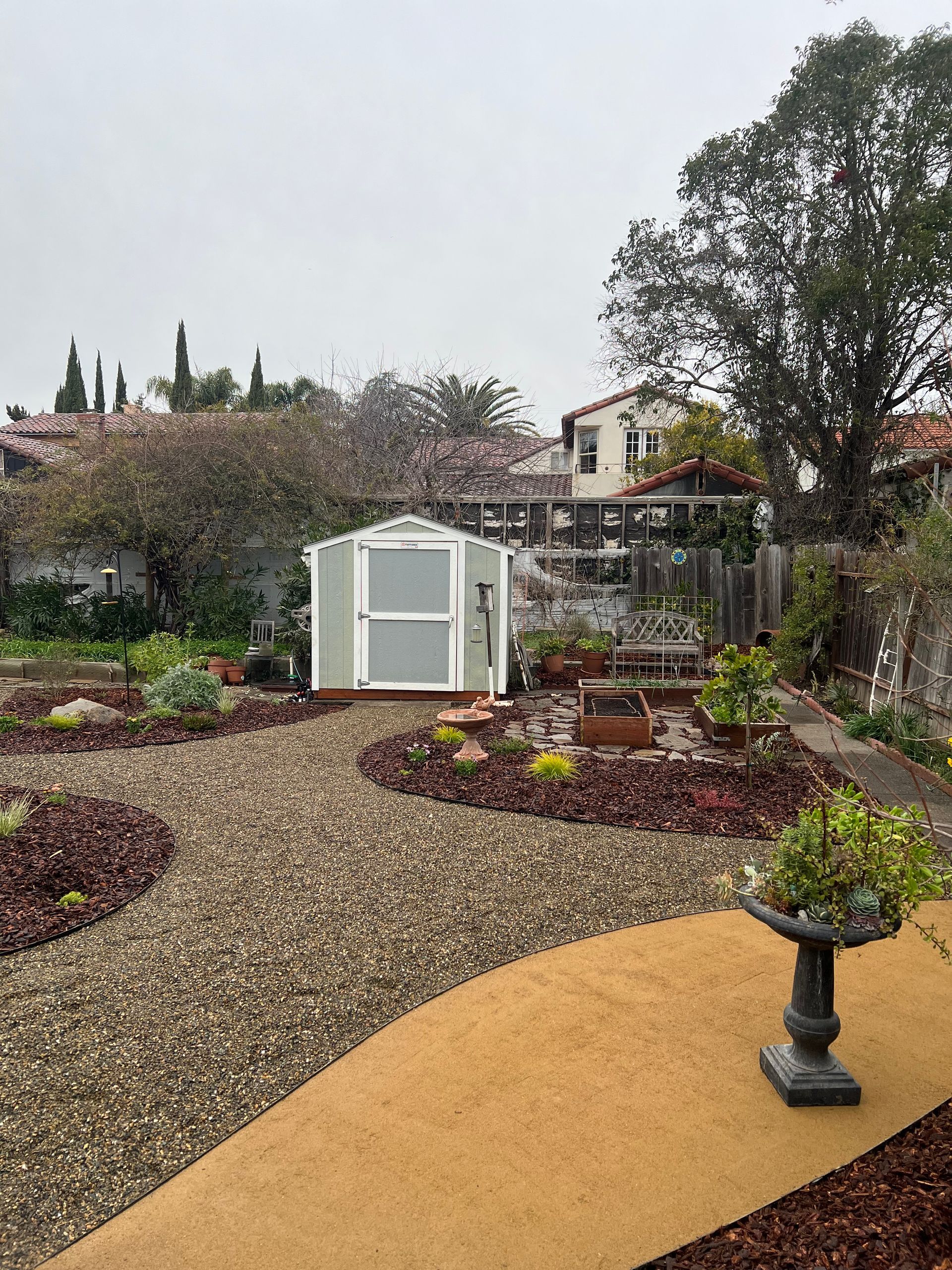 A backyard garden with a shed. Gravel pathway leads to raised beds. Green plants, trees, and a bird bath.