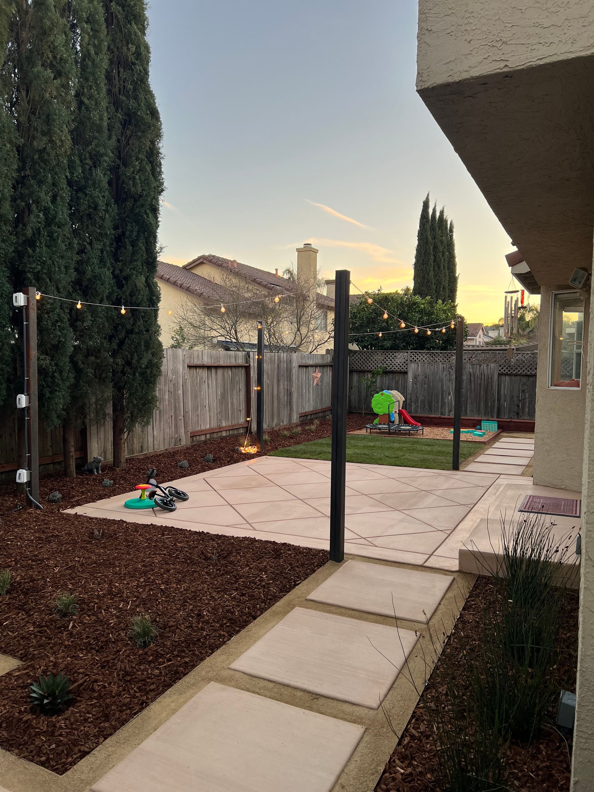 Backyard with concrete patio, lawn, play set, wooden fence, and tall trees at dusk.