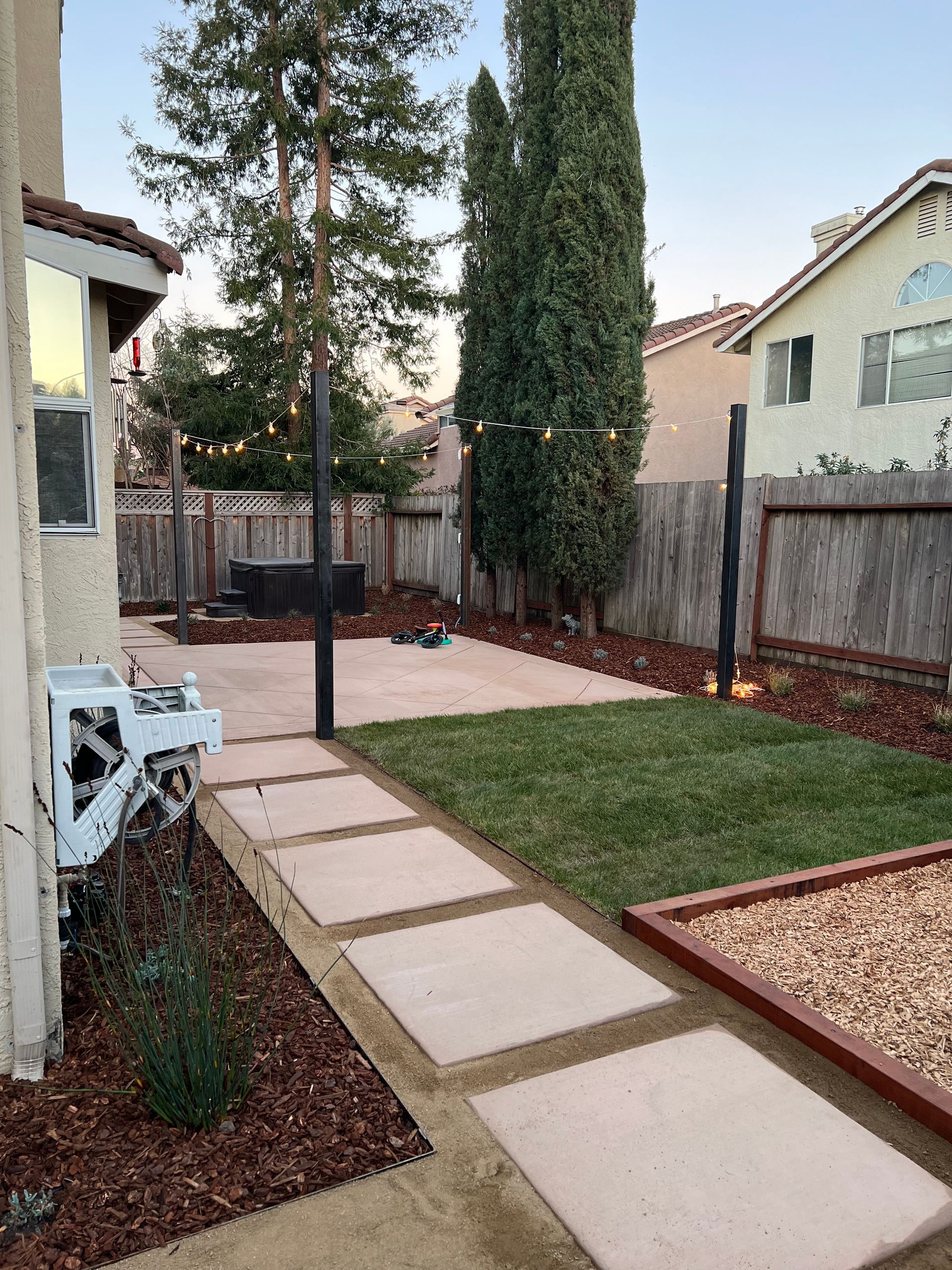 Backyard with concrete path, lawn, gravel area, trees, and a fence.