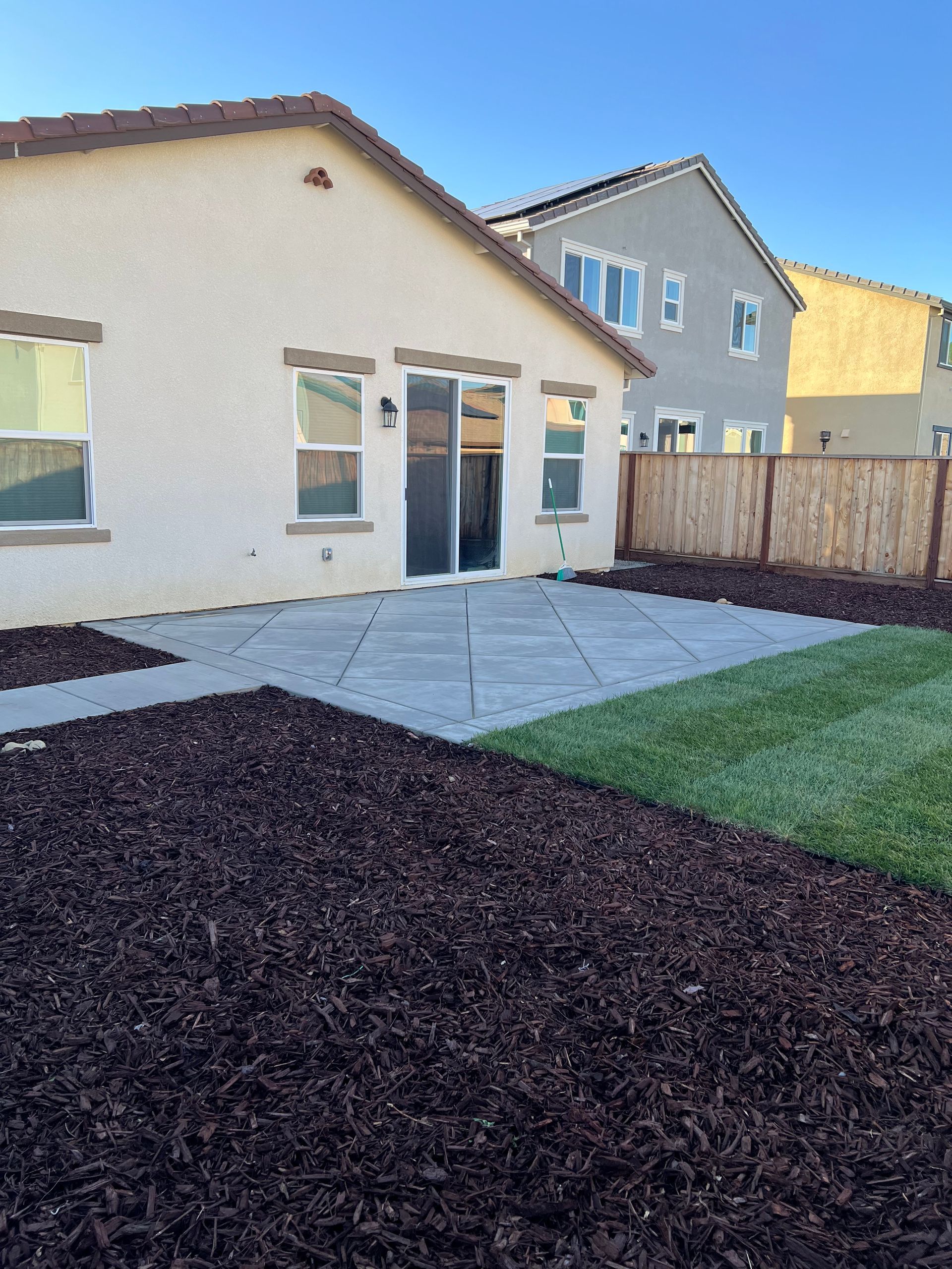 Backyard with patio, grass, and mulch. Tan house with sliding glass door, wooden fence, and blue sky.