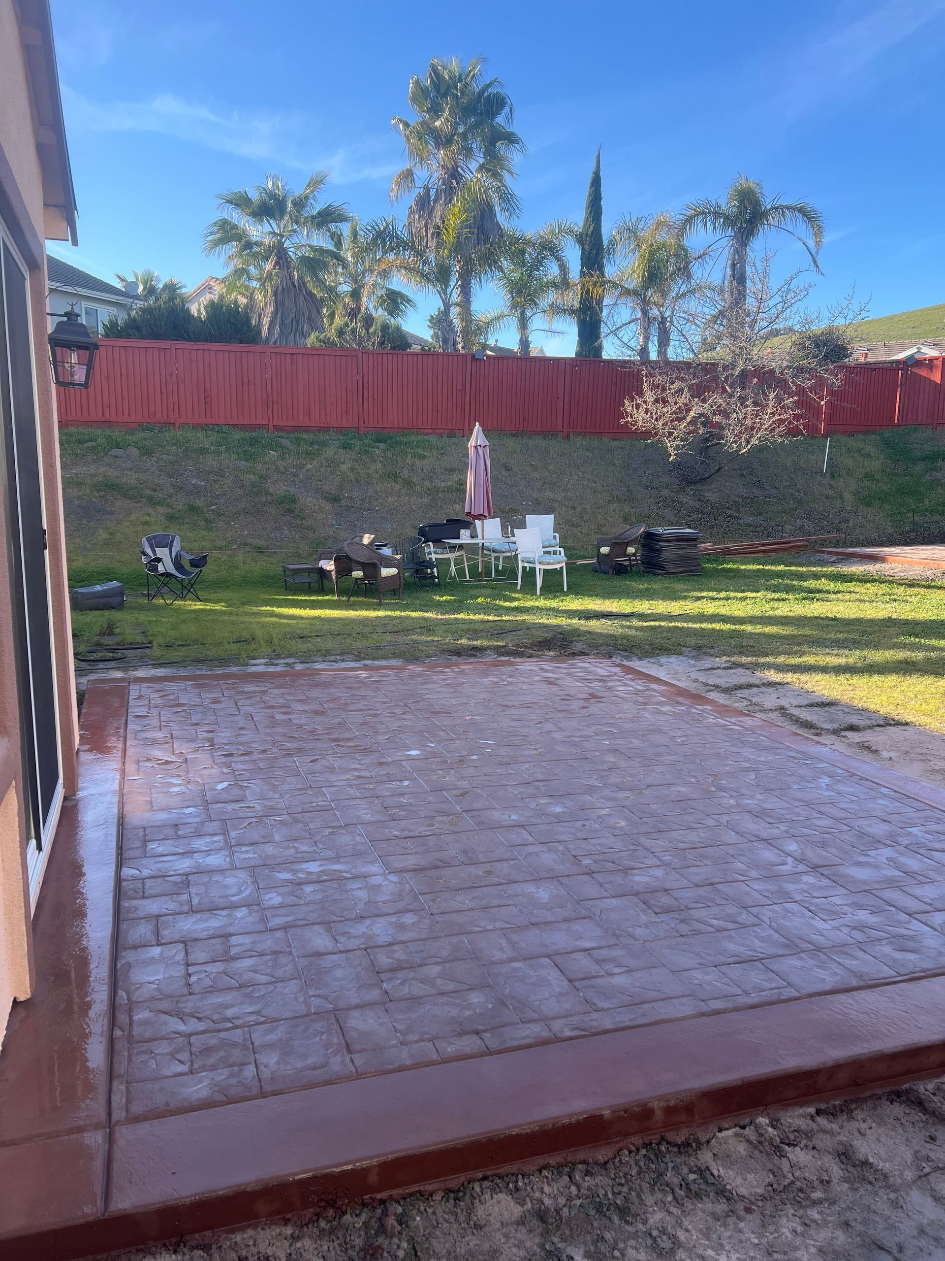 Patio with brick-patterned paving, leading to a backyard with furniture on a grassy area and a red fence.