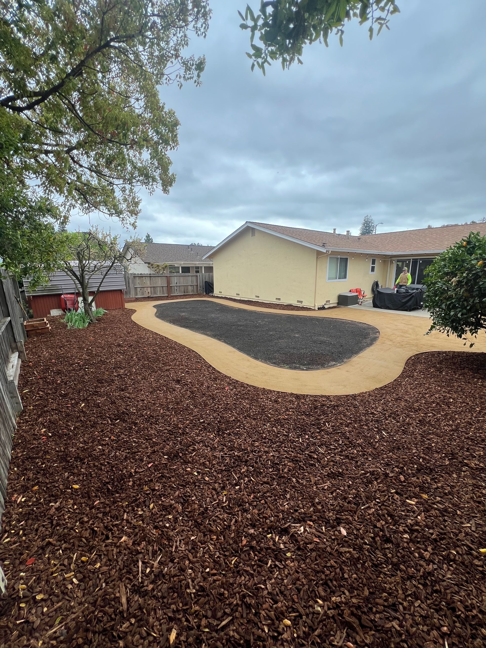 Backyard with wood chips, stone, and a house under a cloudy sky.