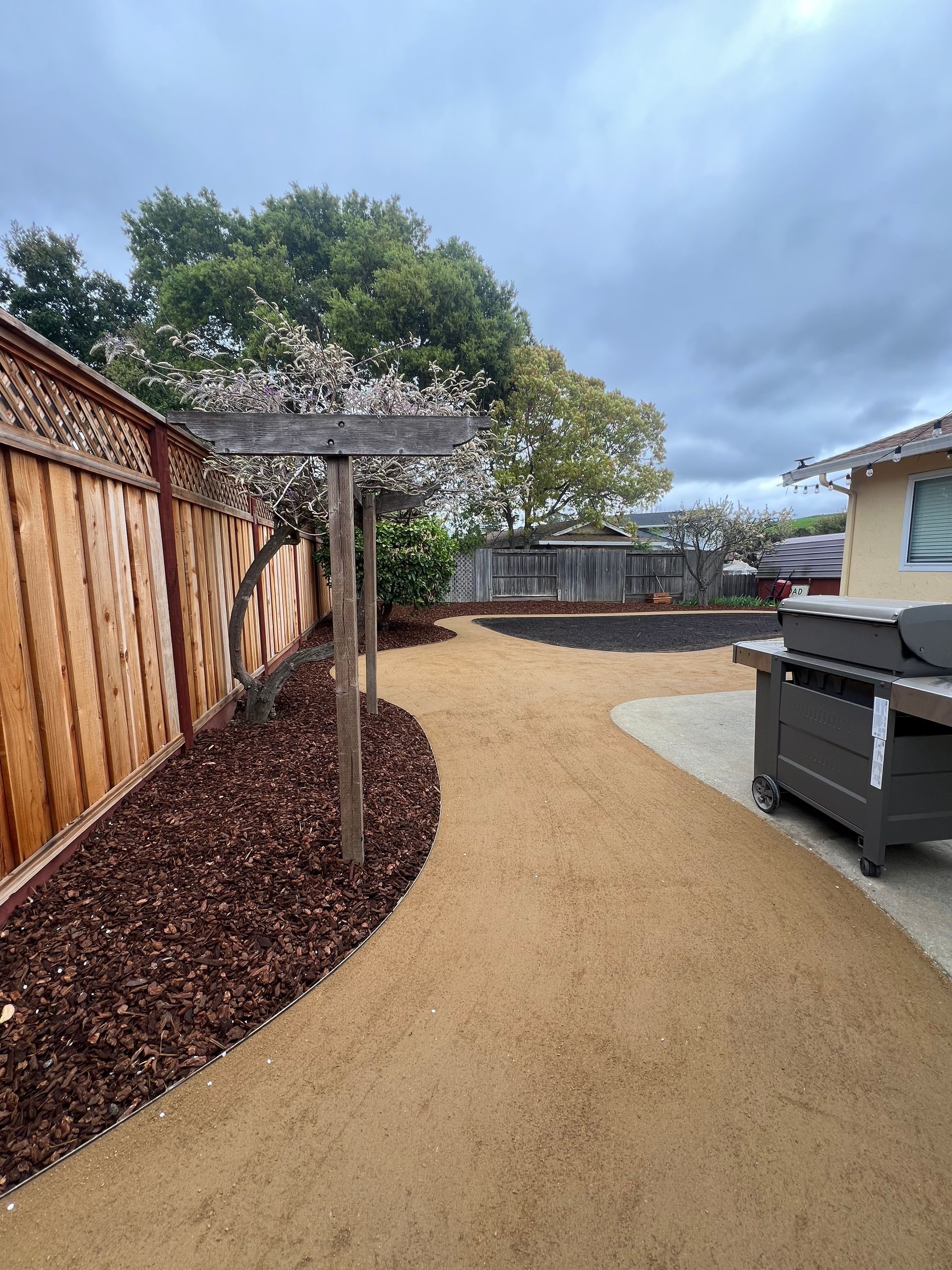 A backyard with a bark mulch bed next to a wooden fence, a gravel path, and a pergola.
