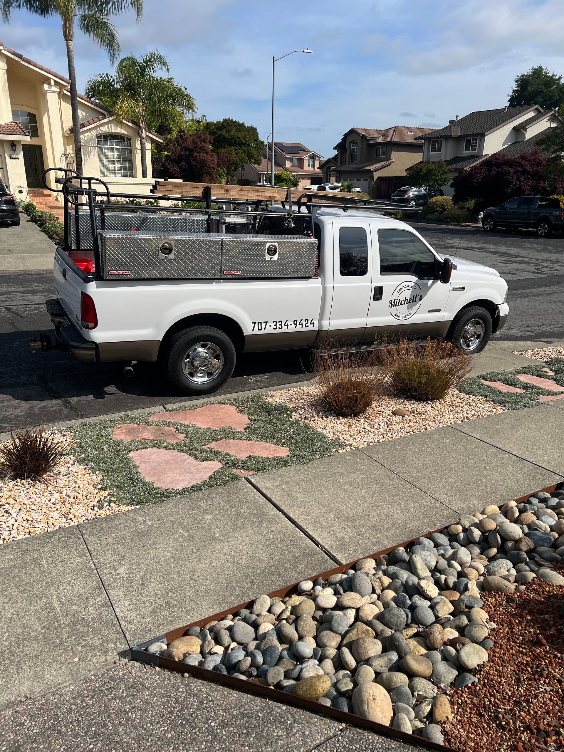 White Ford pickup truck parked on a suburban street. It has a toolbox, ladder rack, and company logo. Sunny day.