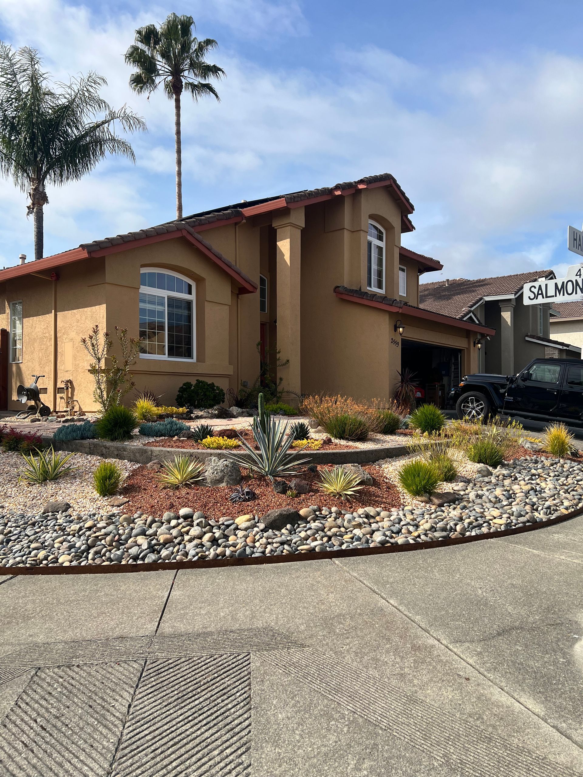 Tan house with red tile roof, lush landscaping, and palm trees on a sunny day.