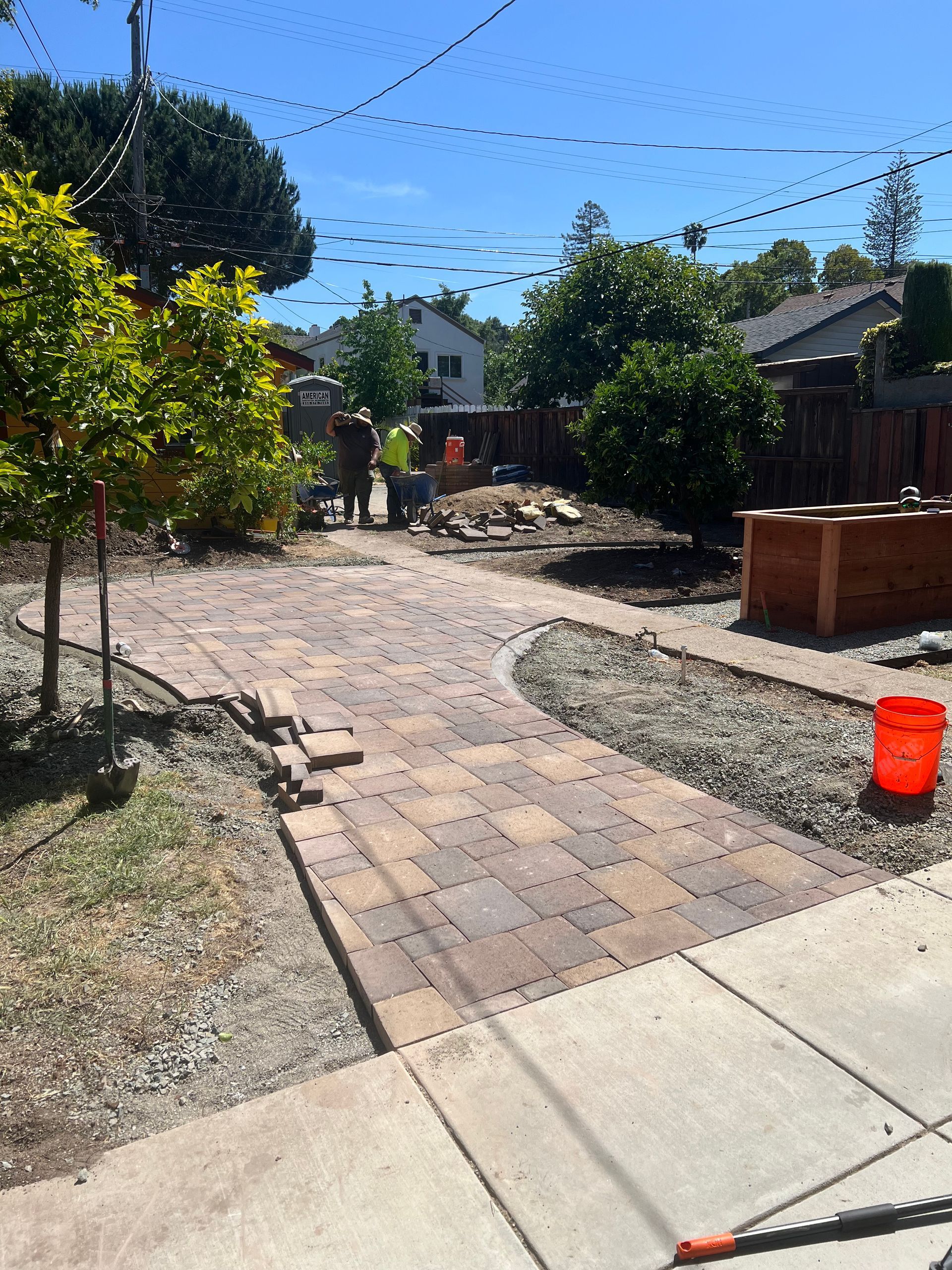 Brick pathway being laid in a yard, workers in the background. Bright sunny day.