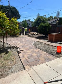 Brick pathway under construction, with workers and materials visible in a sunny yard.