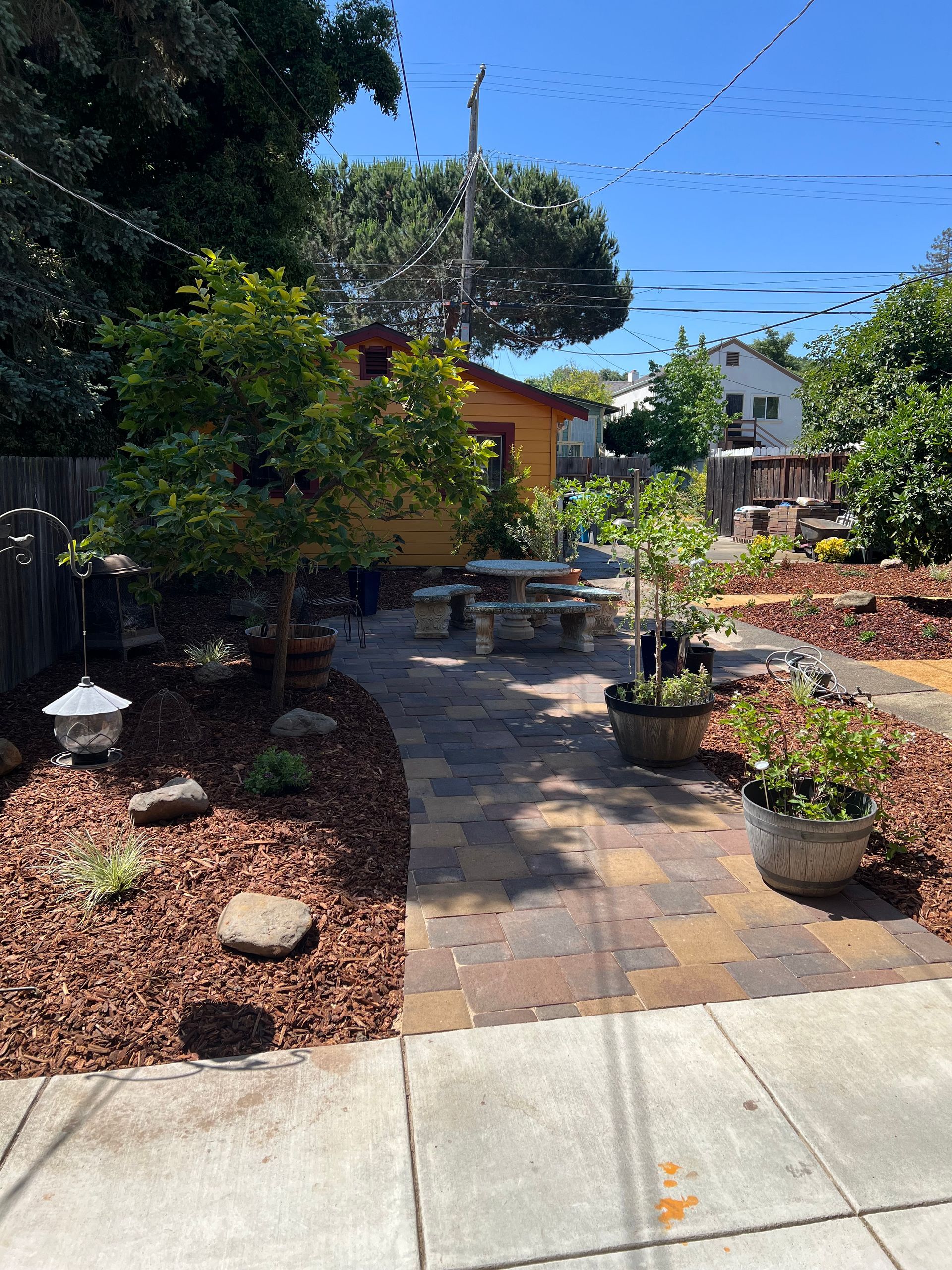 A backyard with a brick walkway, flowerbeds, trees, and a yellow building under a bright blue sky.