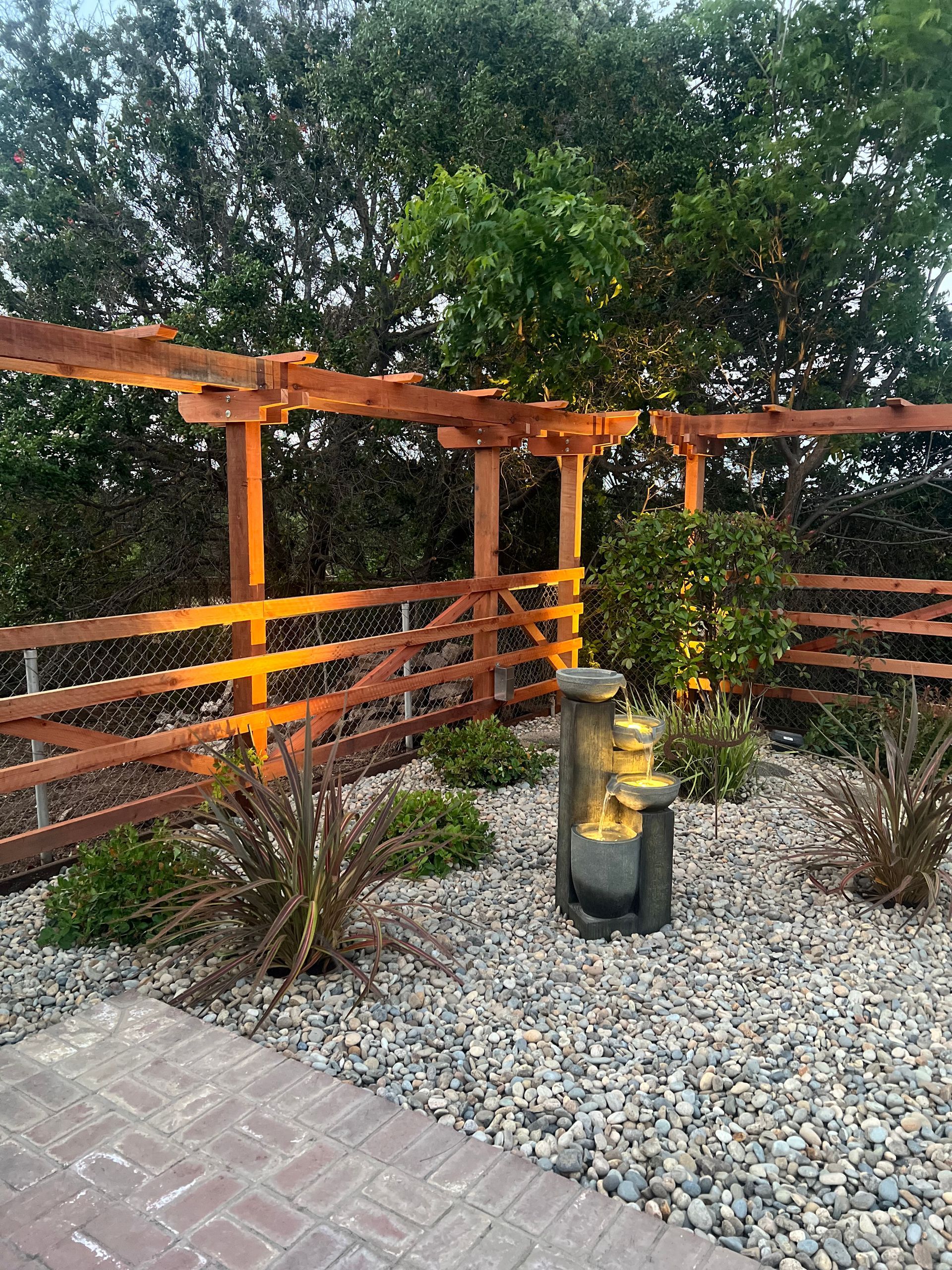 A small garden with a wooden fence, pergola, and a stone fountain, lit with warm light.