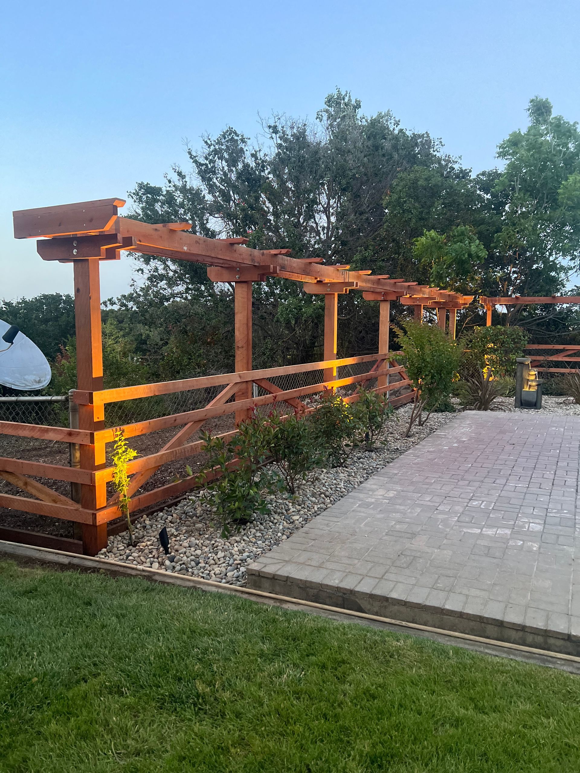 Wooden pergola and fence with brick patio, grass, and trees, bathed in warm sunlight.