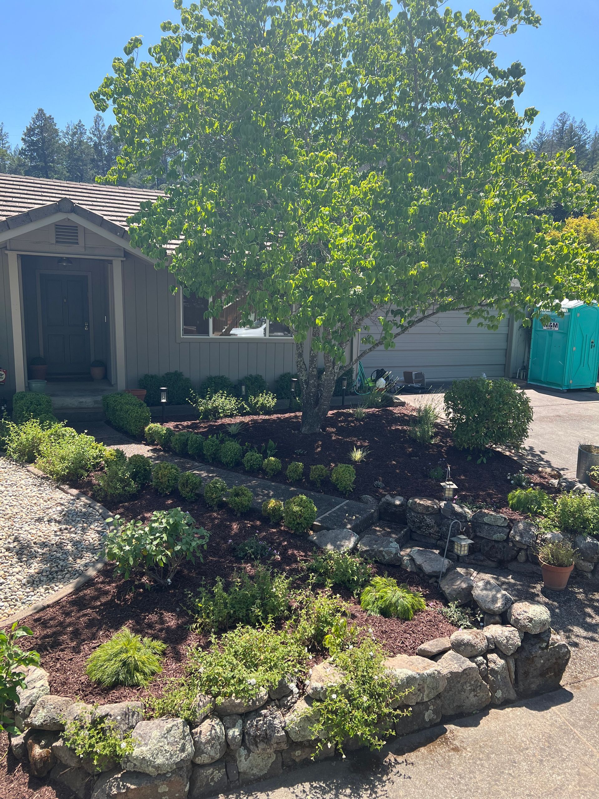House with landscaped yard, a large tree, and a stone retaining wall. Sunny day.