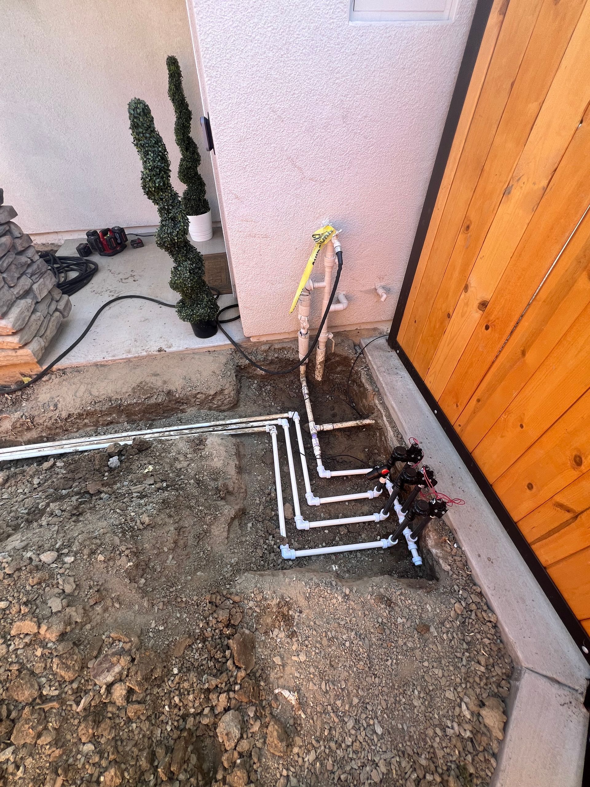 Irrigation system with white pipes branching out from a wall, near a wooden fence and brown dirt.