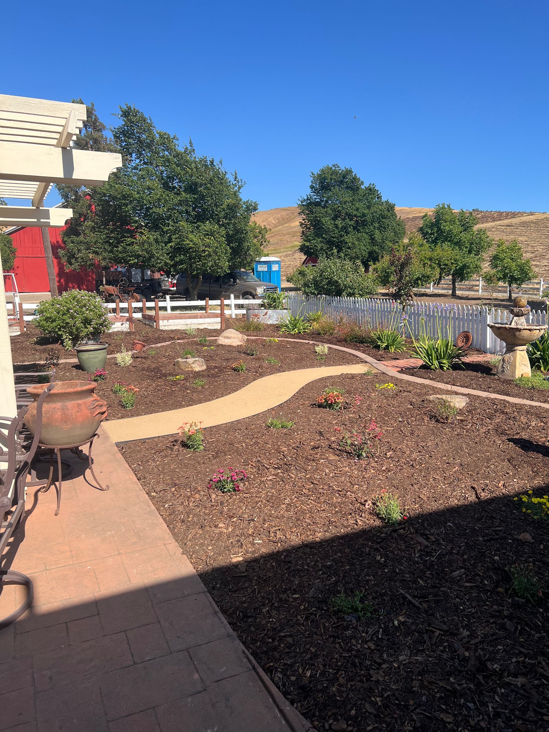 Garden with a mulch pathway, flowers, trees, and a white picket fence under a blue sky.