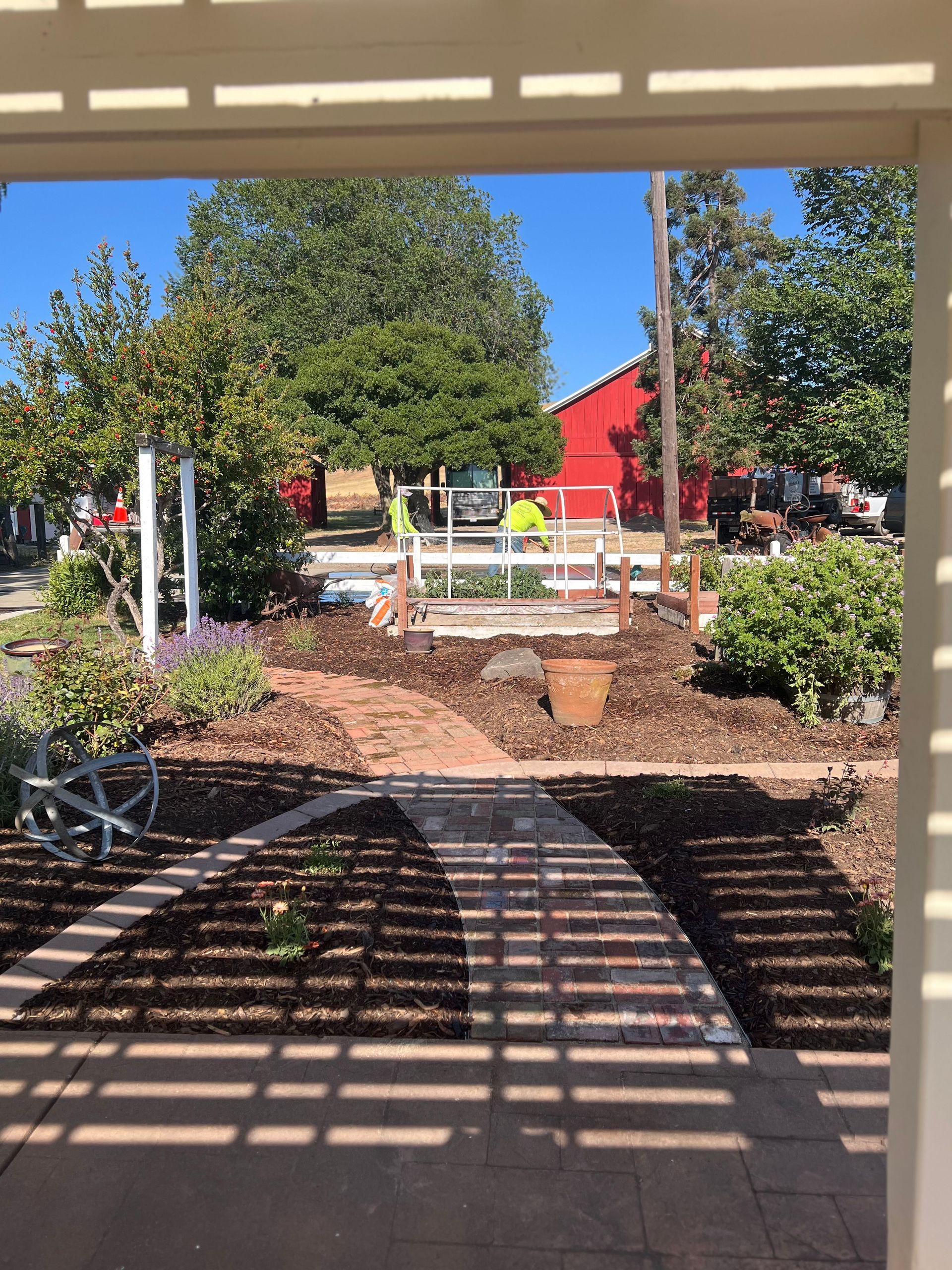 A brick pathway leads to a garden with a red barn in the background, viewed from a porch.