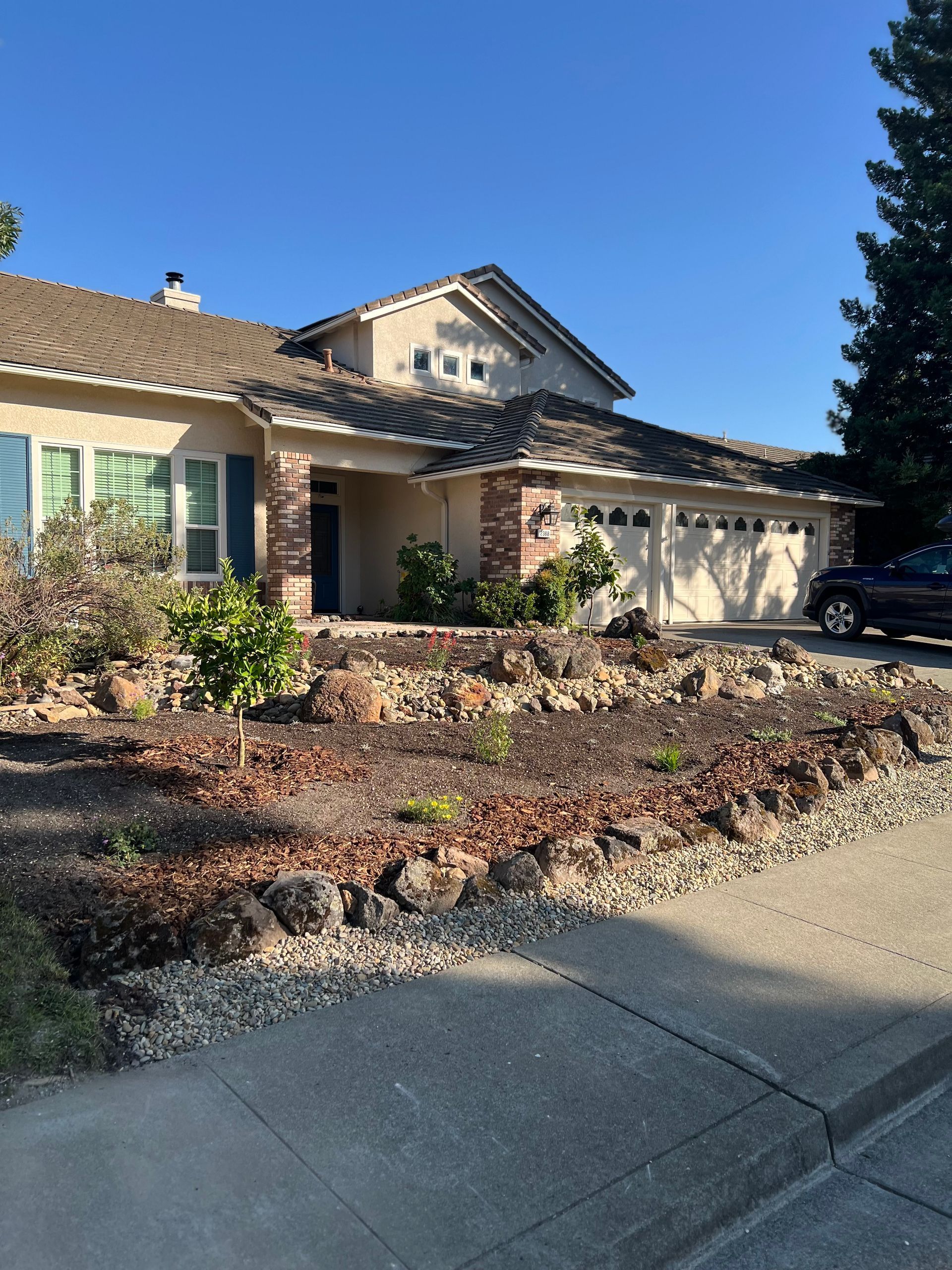 Tan house with brown roof, brick accents, and dry-landscape front yard. Blue sky and parked car.