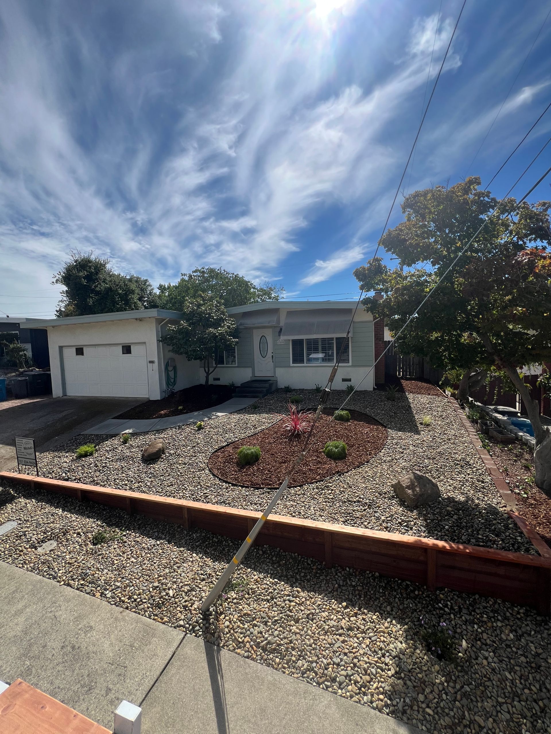 A one-story house with a gravel yard, a garage, and trees under a cloudy sky.